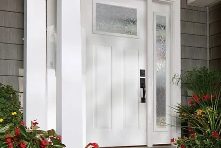 White front door with decorative glass panels surrounded by gray siding and colorful landscaping.